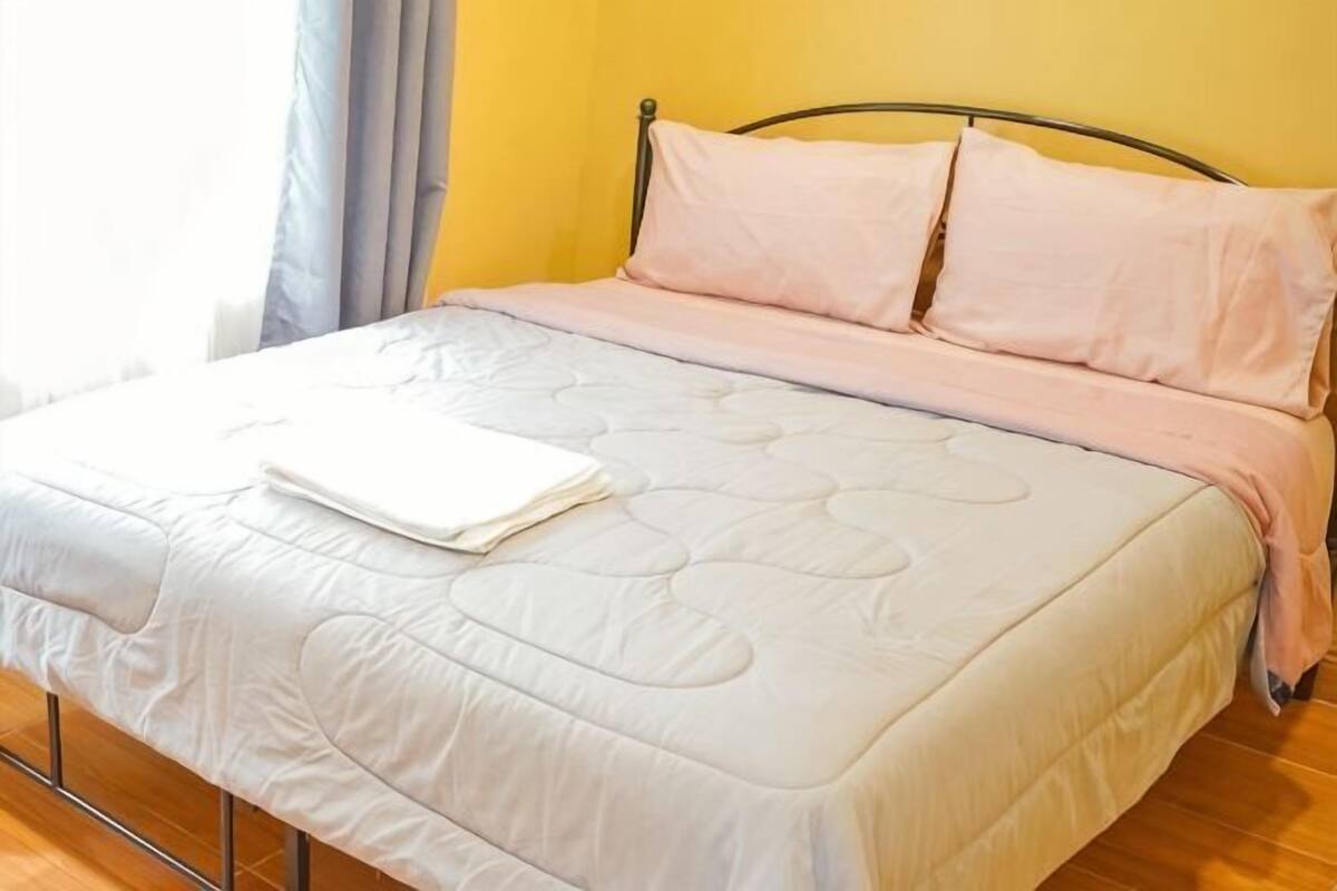 Een bed met een witte sprei, roze kussens, een metalen hoofdbord en een raam met gordijnen.