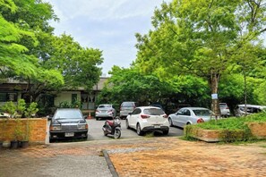 Un espace de stationnement où sont garées plusieurs voitures et une moto, entouré d’arbres et bordé d’un chemin en briques.