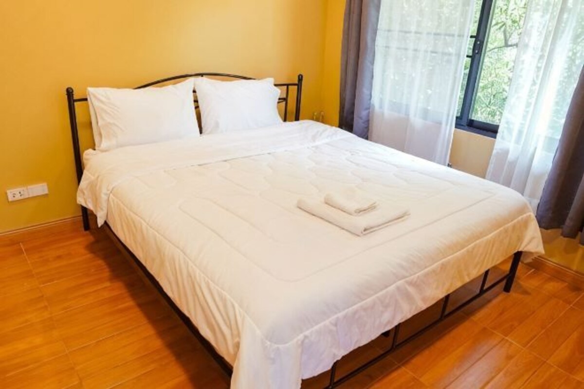 Un lit bien fait, avec du linge de lit blanc et une serviette pliée, une fenêtre avec des rideaux et un plancher en bois.