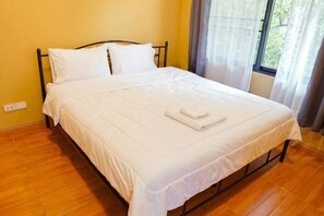 A neatly made bed with white bedding and a folded towel, a window with curtains, and a wooden floor. - Ping Lanna Villa (Chiang Mai)