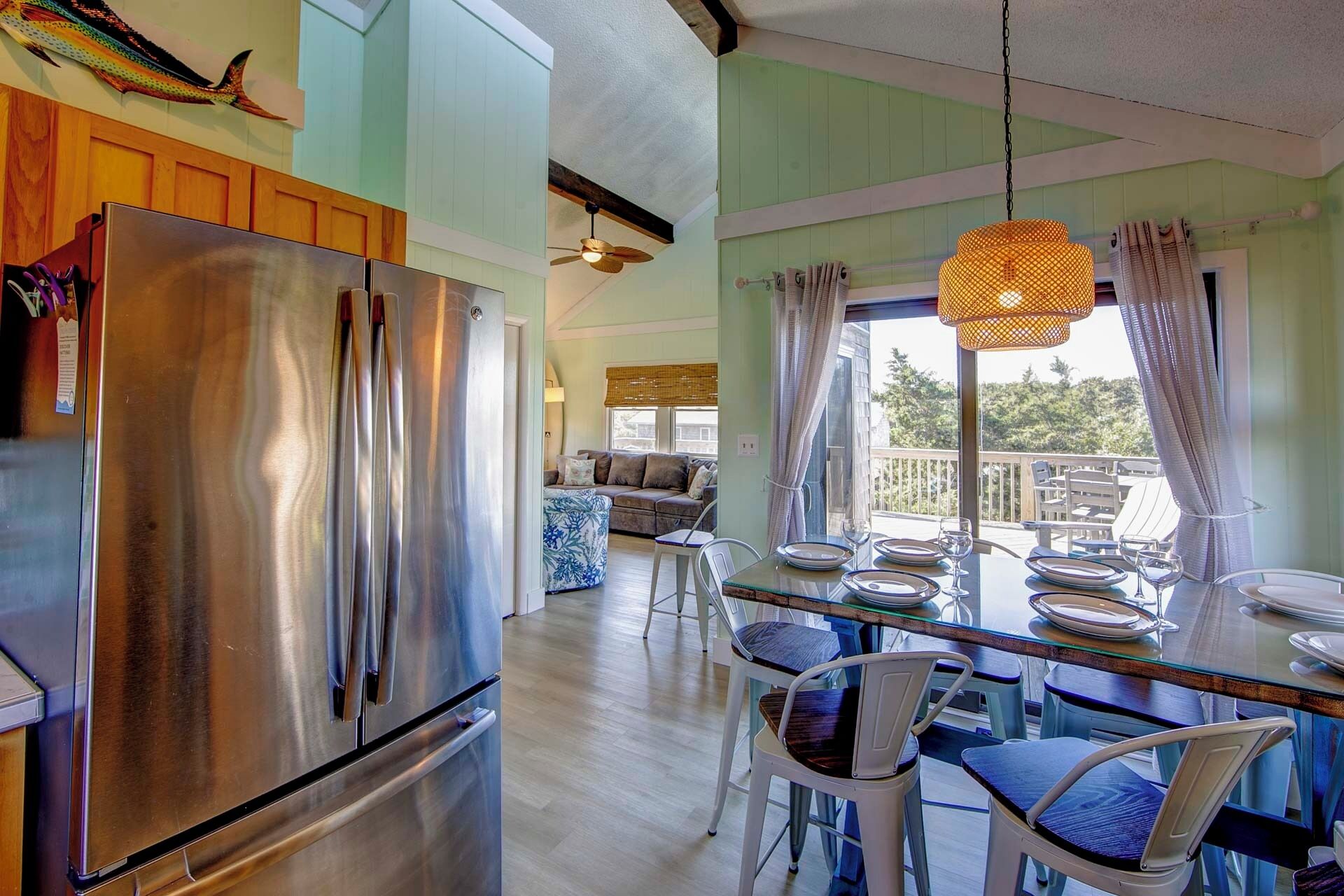 A modern kitchen with a stainless steel refrigerator, a dining area with a glass table and chairs, and a view of the outdoors through large windows.