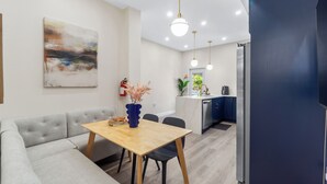 Ein modernes Wohnzimmer mit einem grauen Sofa, einem Holzesstisch und einer blauen Vase. Im Hintergrund ist eine Küche mit Kühlschrank und Spüle zu sehen.