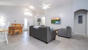 Un amplio espacio de sala con un sofá gris, una mesa de comedor con sillas, un ventilador de techo y un cuadro en la pared.