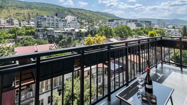 Ein Balkon mit Tisch, einer Weinflasche und einem Glas. Der Balkon bietet Blick auf eine Stadtlandschaft mit Gebäuden und einer Hügelkulisse im Hintergrund.
