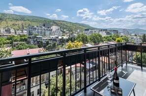 Un balcon avec une table, une bouteille de vin et un verre. Le balcon offre une vue sur le paysage urbain, avec des bâtiments et une colline en arrière-plan.