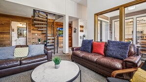 Una sala de estar moderna con un sofá de cuero marrón, una mesa redonda de centro con una planta, y una escalera de madera.