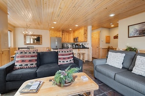 En moderne stue med en sort sofa og en grå sofa, begge med mønstrede puder. Et træsofabord med en plante i midten. Værelset har trælofter og et køkken i baggrunden.