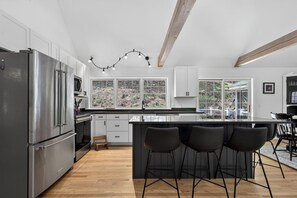 A modern kitchen with a stainless steel refrigerator, a microwave, a large window, and a bar area with stools. - Spacious 5-bedroom estate in charming Ogunquit with AC and 700 square foot deck. (Ogunquit)