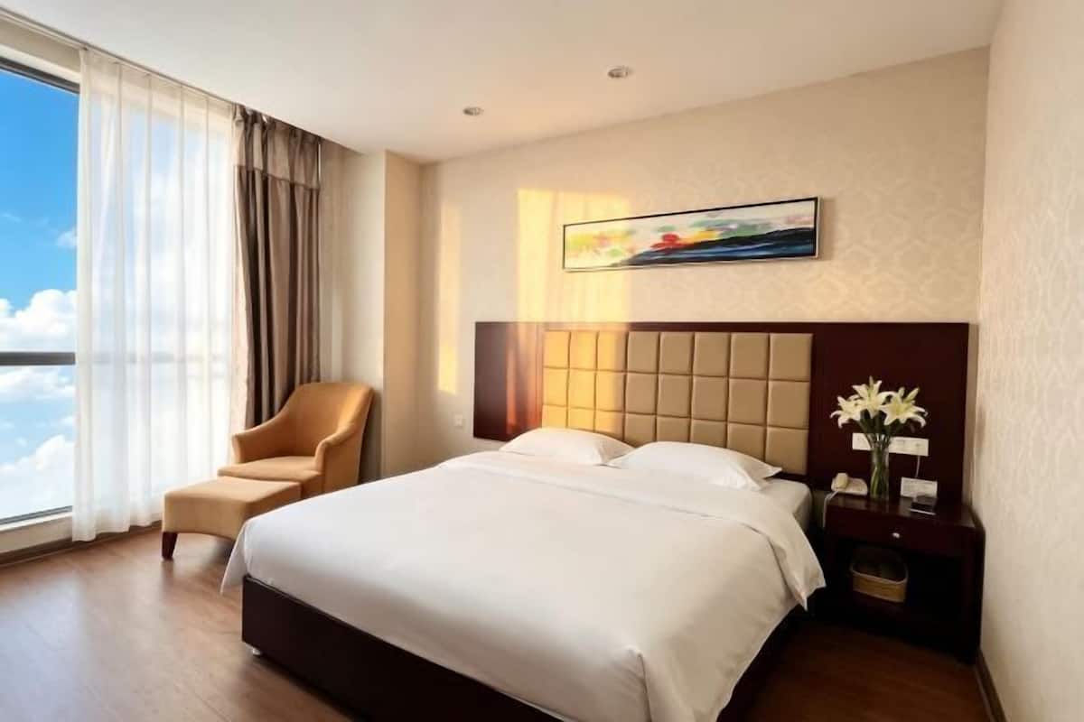 Hotellihuone, jossa on suuri sänky, tuoli, yöpöytä, jossa on maljakko täynnä kukkia, ja taulu seinällä.