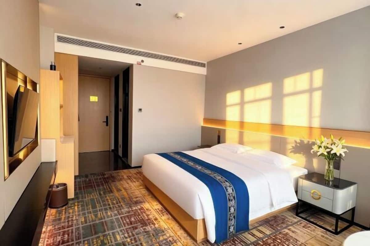A hotel room with a bed, a bedside table with a vase, a television, and a patterned carpet.