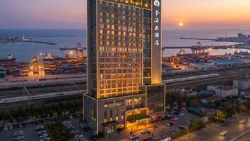 A high-rise hotel with multiple floors, a parking lot, and a view of the sea and port facilities.
