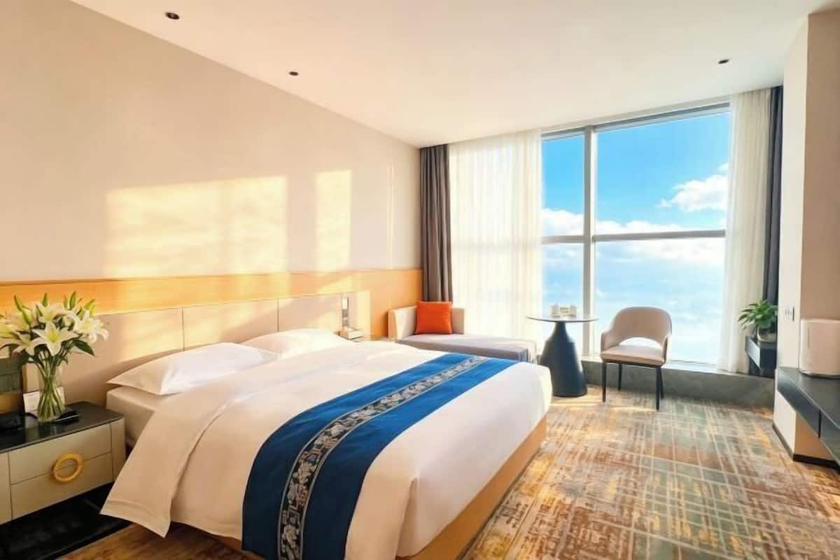 A hotel room with a large bed, a desk, a chair, and a view of the sky.