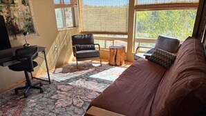 Un salón acogedor con un sofá marrón, una silla de escritorio negra y una mesa de centro de madera. Un ventanal ofrece vistas a árboles fuera.