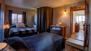 Un dormitorio con dos camas, una mesita de noche, un aparador, una ventana con cortinas y una puerta que da a otra habitación.