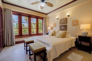 Une chambre d’hôtel avec un grand lit, deux tables de chevet, un ventilateur de plafond et une vue sur les palmiers par les fenêtres.