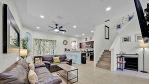 A modern living room with a brown leather sofa, a coffee table, and a ceiling fan. A kitchen is visible in the background with a microwave and cabinets. - Near Disney Regal Palms Renovated Resort-New Pool-Lazy River-Arcade-Kid Friendly (Davenport)