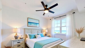 A bedroom with a bed, two bedside tables, a ceiling fan, a window with curtains, a wall-mounted painting, and a wicker basket. - Enjoy a luxury 2BR/2BA Condo in beautiful Ave Maria Golf and Country Club (Ave Maria)