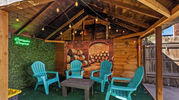 An outdoor seating area with wooden tables and chairs, a neon sign, and a mural of barrels.