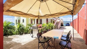 Un patio al aire libre con una mesa y sillas bajo un gran paraguas, una parrilla, y una vista de un edificio y un automóvil al fondo.
