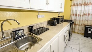 Una cocina con armarios blancos, un lavamanos de acero inoxidable, un tostador, un hervidor y un cubo de basura.