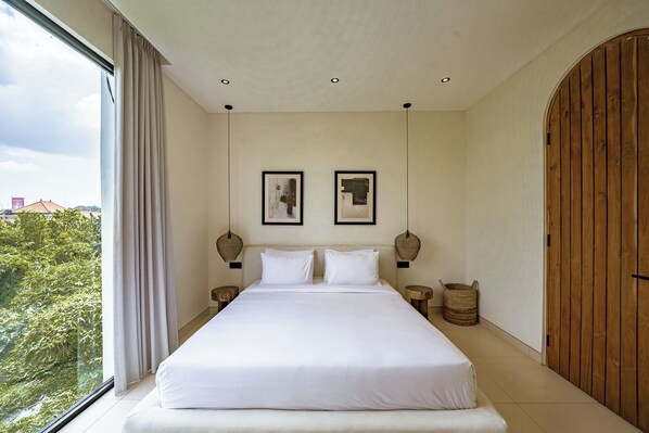 A bedroom with a large bed, two framed pictures on the wall, a wooden door, and a view of greenery outside. - Villa JB Canggu (Canggu)