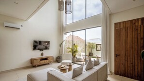 A modern living room with a large sofa, a TV mounted on the wall, and a wooden cabinet. There is a large window providing a view of the outside. - Villa JB Canggu (Canggu)