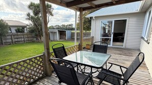 A covered patio with a table and chairs, a wooden deck, and a view of a house and yard. - Kowhai on Douglas – Your Relaxing Escape (Levin)