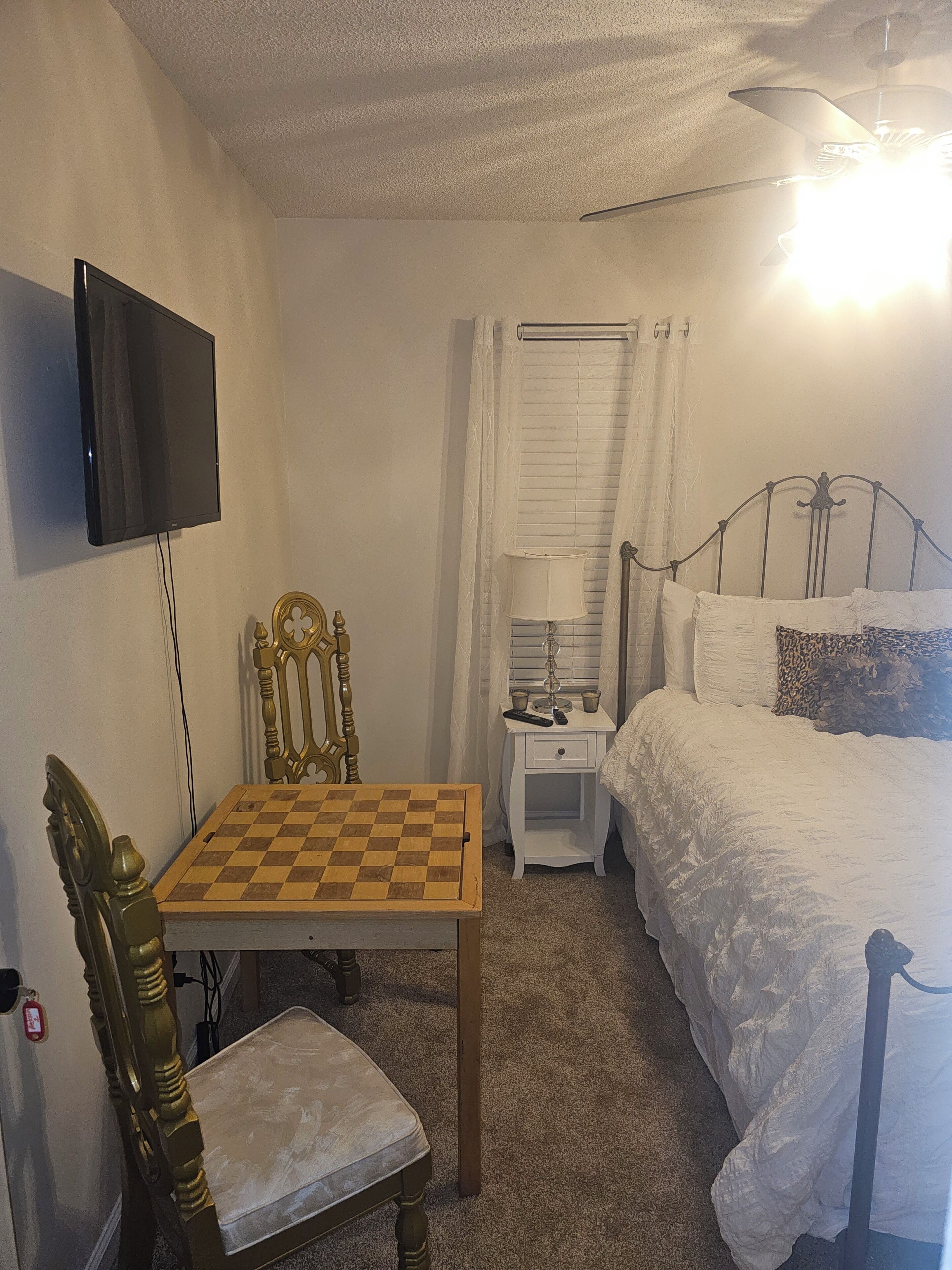 Ein Schlafzimmer mit Bett, Schreibtisch mit Schachbrett, Wandfernseher und Deckenventilator.