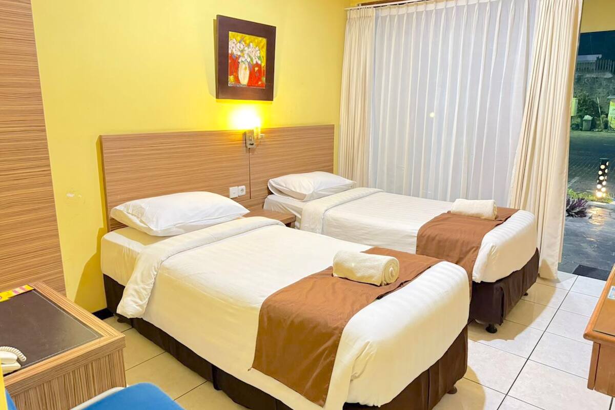 Une chambre d’hôtel avec deux lits, une table de chevet, une chaise, un tableau au mur et une fenêtre avec des rideaux.