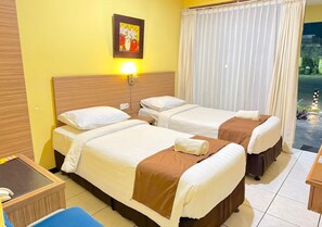 Une chambre d’hôtel avec deux lits, une table de chevet, une chaise, un tableau au mur et une fenêtre avec des rideaux.