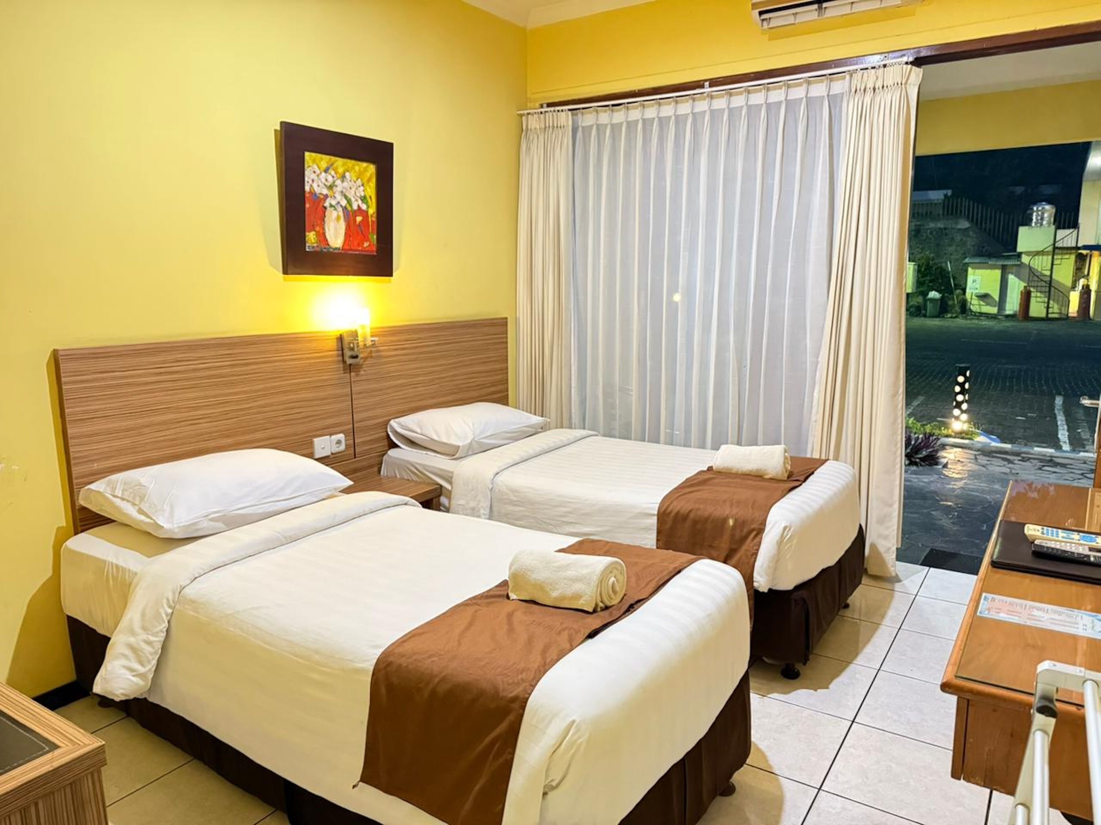 A hotel room with two beds, a nightstand, a lamp, a painting, and a view of the outside.