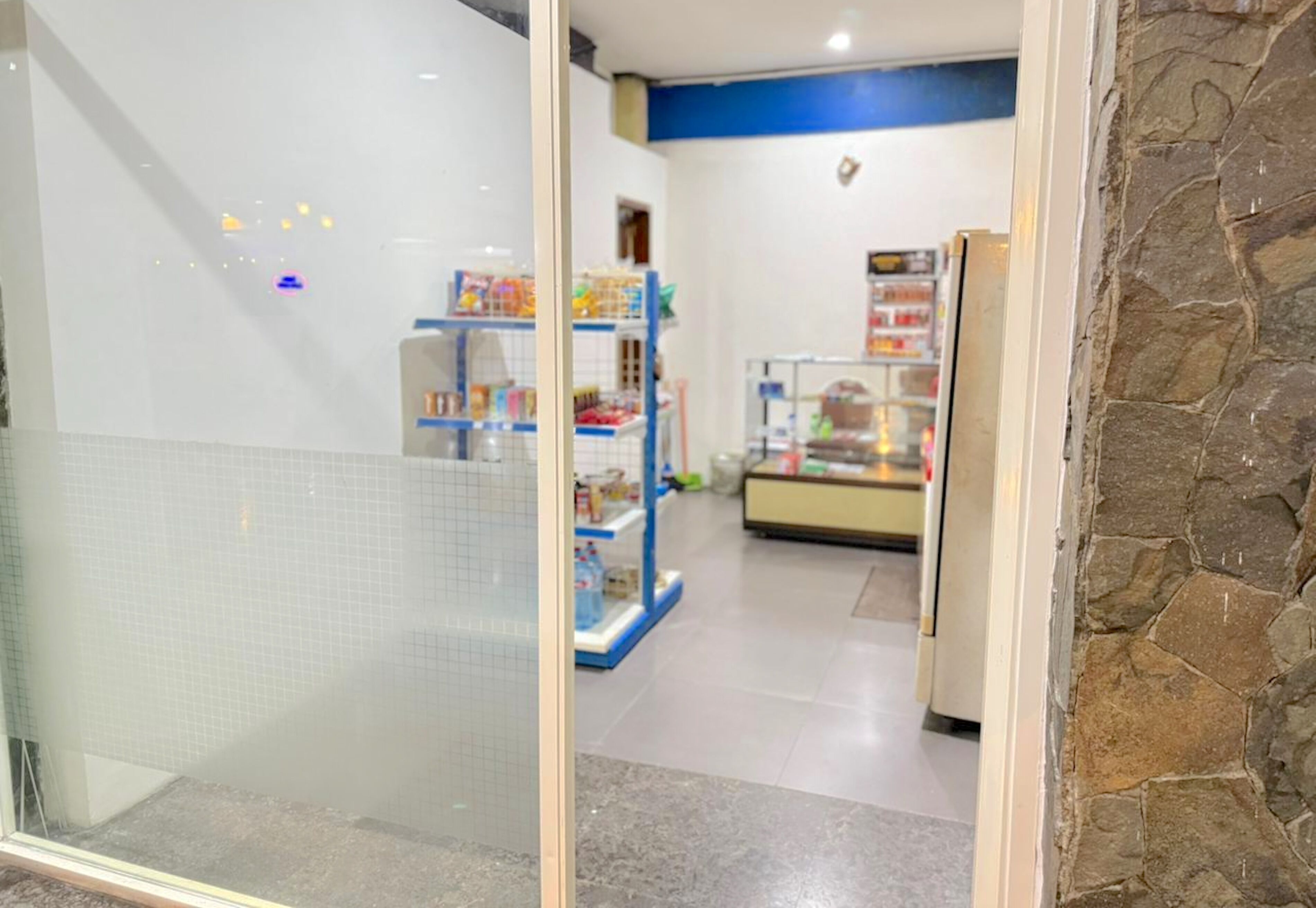 Um corredor com parede revestida de azulejos, parede de pedra à direita e prateleiras com diversos objetos ao fundo.