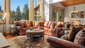 A spacious living room with a large leather sectional sofa, a round coffee table, and a view of a forest through large windows. - Lakota Dreamcatcher North | Sauna | Slope Views | Private Hot Tub (Winter Park)