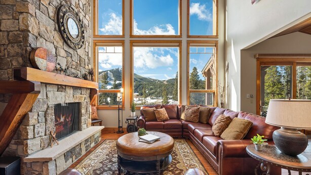A cozy living room with a stone fireplace, leather furniture, and large windows offering mountain views. - Lakota Dreamcatcher North | Sauna | Slope Views | Private Hot Tub (Winter Park)