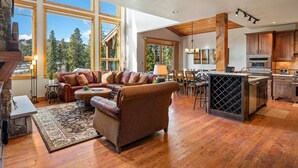 A spacious living area with a large sofa, a dining table, and a kitchen with wooden cabinets and a wine rack. - Lakota Dreamcatcher North | Sauna | Slope Views | Private Hot Tub (Winter Park)