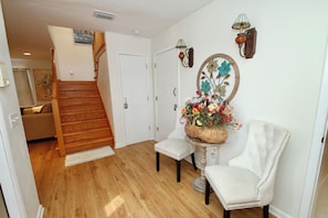 A hallway with a wooden staircase, a white chair, a small table with a basket of flowers, and wall-mounted lamps.