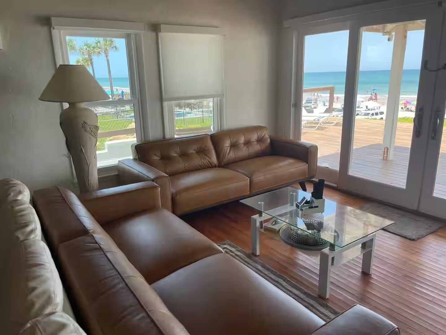 A living room with a brown leather sofa, a glass coffee table, and a lamp. There are large windows with a view of the beach and ocean.