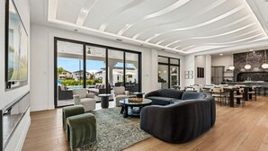 Uma sala de estar ampla com design moderno no teto, uma grande janela com vista para uma piscina e uma cozinha com painel de fundo de pedra.