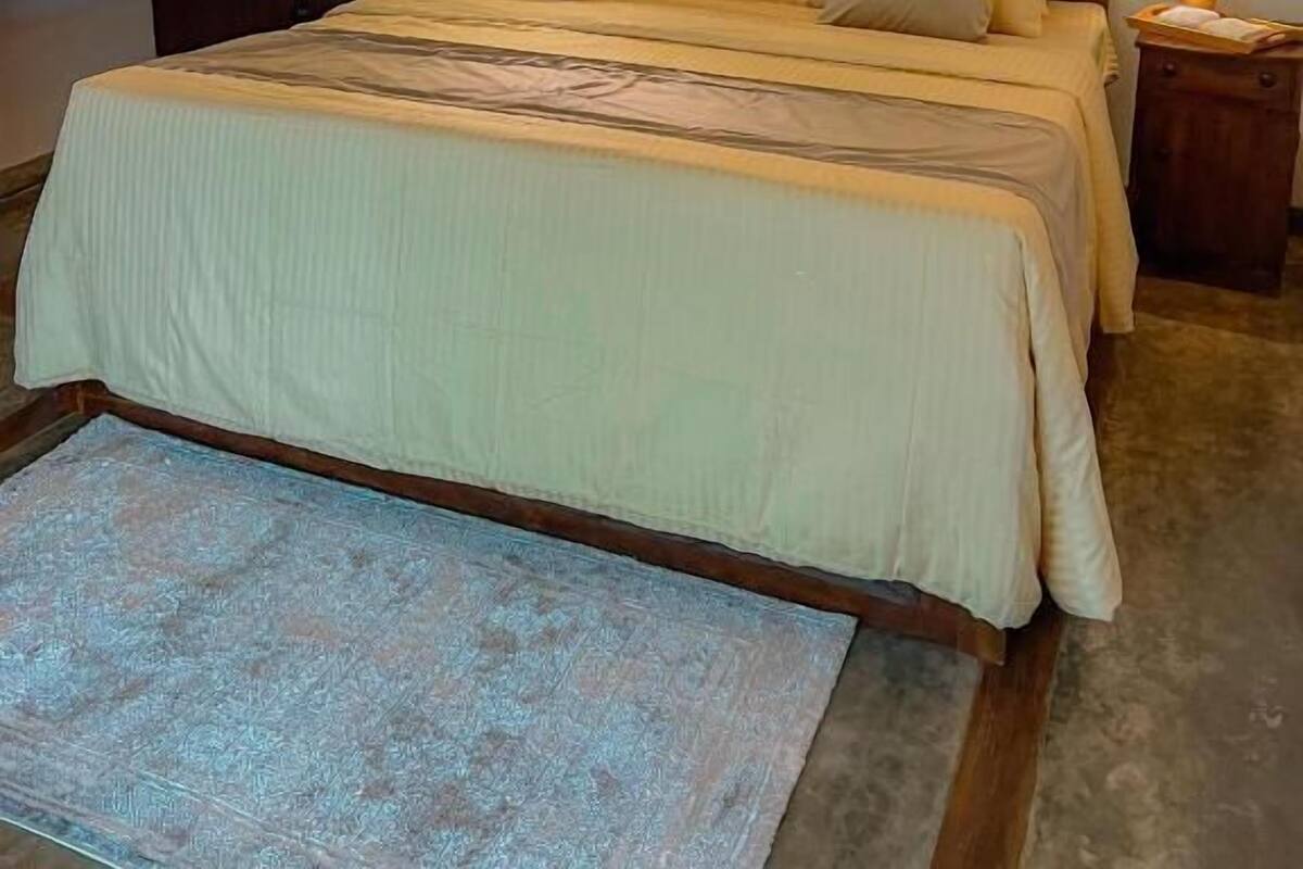Una cama bien tendida con cabecera de madera, una mesita de noche con lámpara y una pequeña planta en maceta.