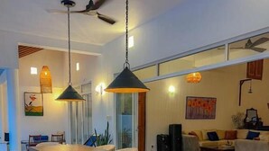 Un espace de salle à manger moderne avec une longue table en bois, des chaises blanches avec des coussins, des luminaires suspendus et un ventilateur de plafond.