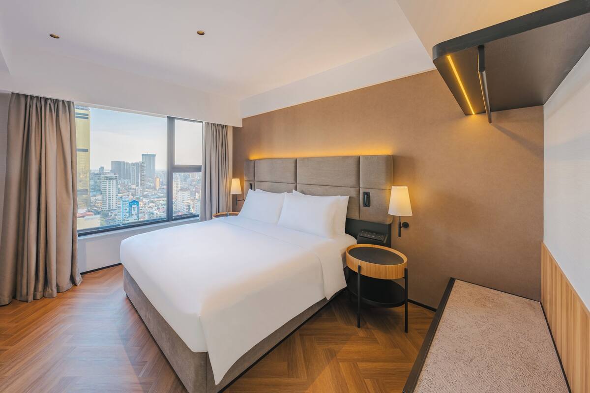 Ein Hotelzimmer mit einem großen Bett, einem Nachttisch, einer Lampe und Blick auf die Stadt.