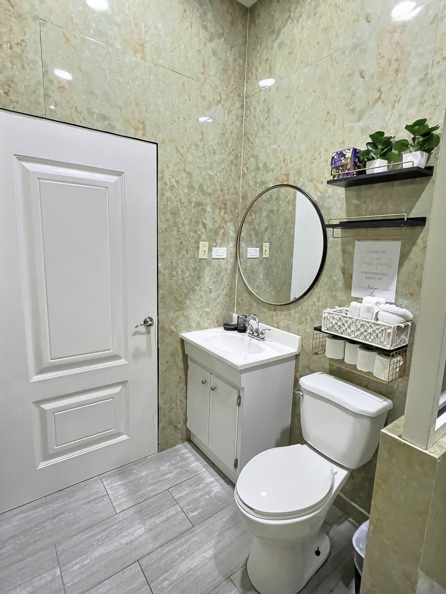A bathroom with a white door, a sink with a mirror above it, a toilet, and a shelf with toiletries.