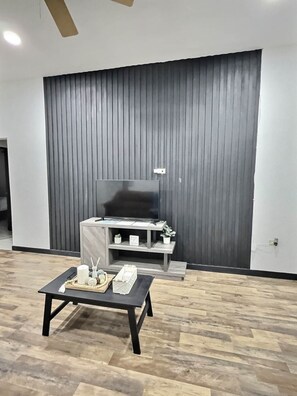 Un moderno salón con un televisor de pantalla plana grande, una mesa de centro de madera y una pared oscura con líneas verticales.