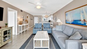 Uma sala de estar espaçosa com uma mesa de centro branca, sofá cinzento e poltronas estampadas. Uma área de jantar visível ao fundo, com uma mesa e cadeiras brancas.