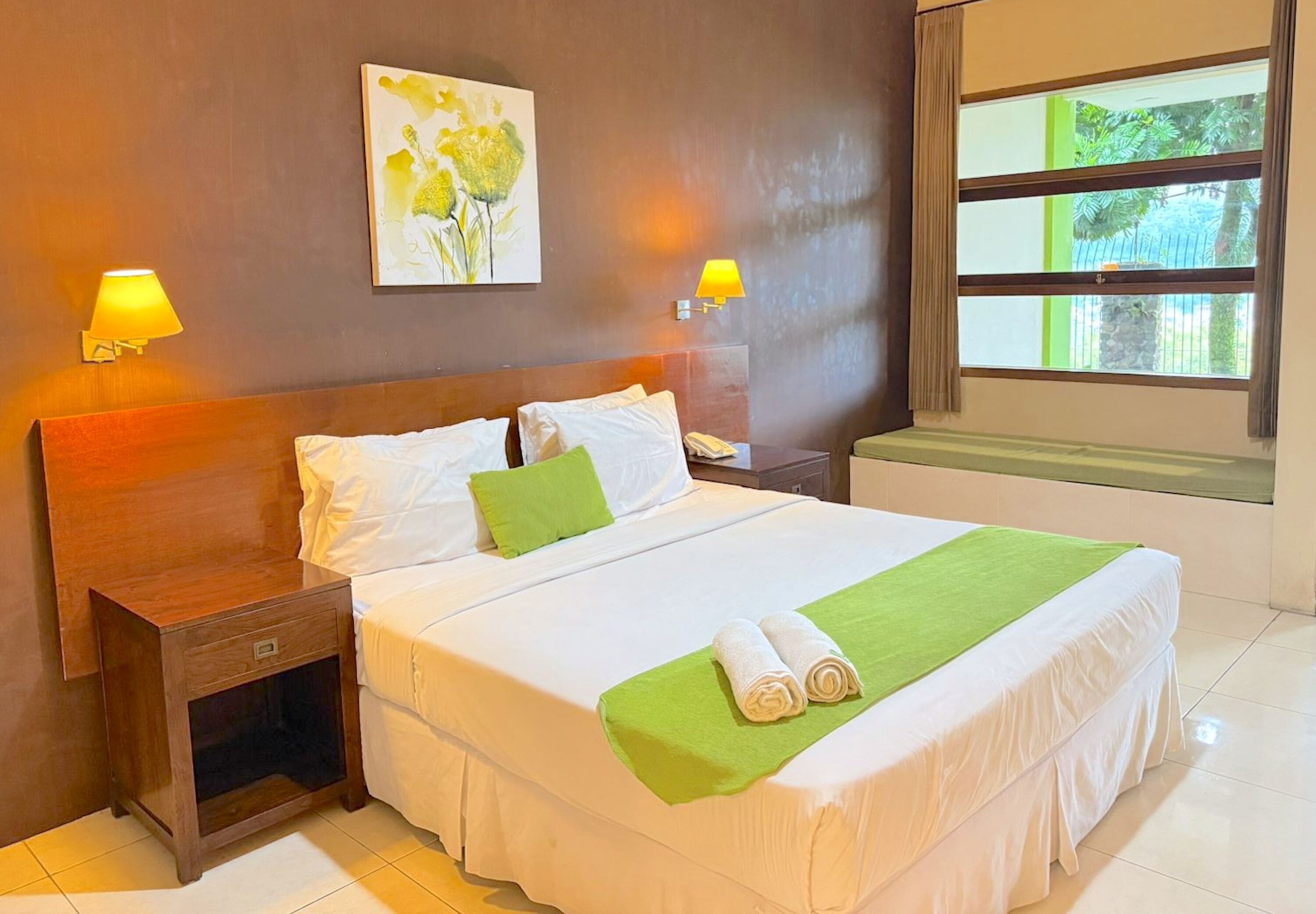 A neatly made bed with a green towel and a white blanket, a wooden nightstand with a lamp, and a painting on the wall.