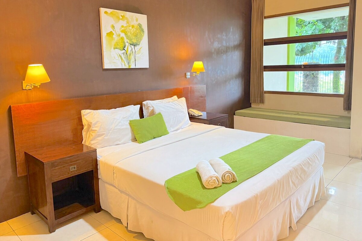 A neatly made bed with a green towel and a white blanket, a wooden nightstand with a lamp, and a painting on the wall.