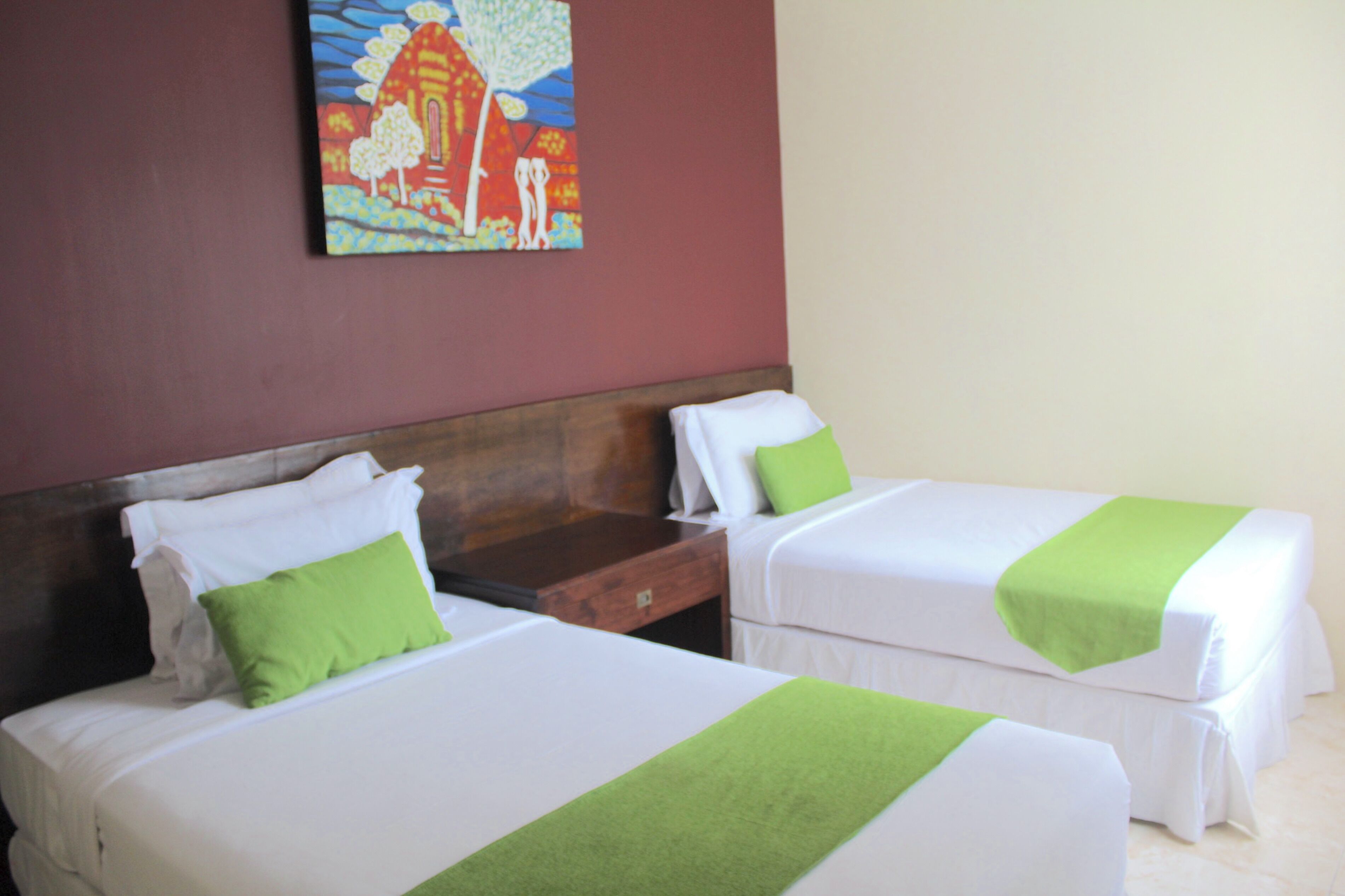 Two beds with white linens and green accents, a wooden headboard, and a colorful painting on the wall.