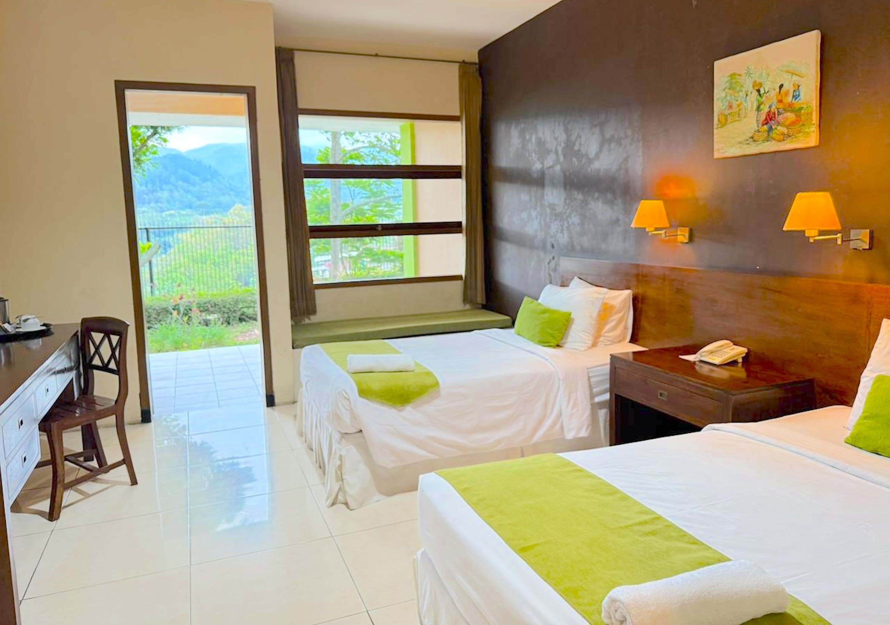 Une chambre d’hôtel avec deux lits, un bureau et une chaise. Il y a une fenêtre donnant sur un paysage verdoyant et un tableau accroché au mur.