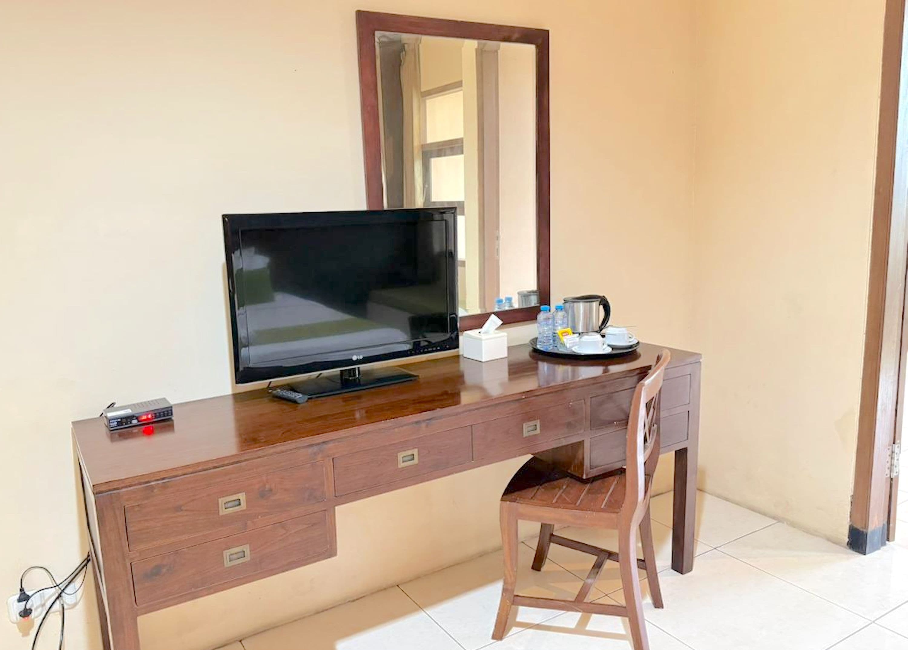 Un escritorio de madera con una televisión de pantalla plana, un espejo, una silla y una bandeja con una cafetera y tazas.