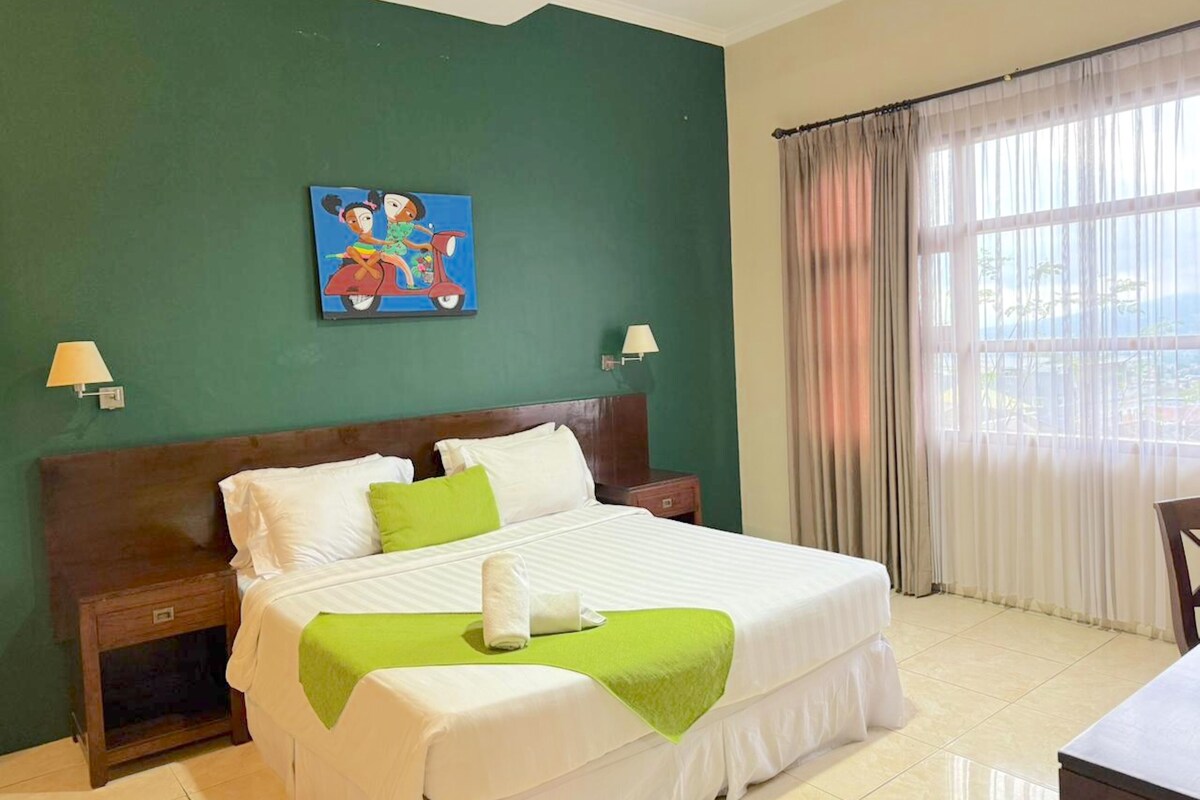 A hotel room with a large bed, green walls, a painting above the bed, two bedside lamps, and a window with curtains.